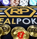 Stacks of poker chips in various colors are arranged in the background, while in the foreground, there are numerous golden coins featuring the Bitcoin symbol. The text 'REALPOKER' is prominently displayed in gold and silver lettering on a dark background.