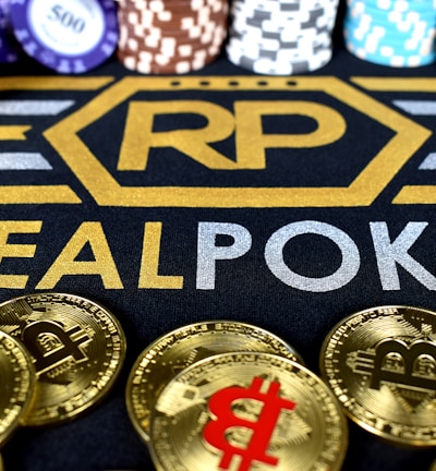 Stacks of poker chips in various colors are arranged in the background, while in the foreground, there are numerous golden coins featuring the Bitcoin symbol. The text 'REALPOKER' is prominently displayed in gold and silver lettering on a dark background.