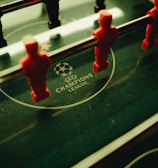 A foosball table is shown with red and black player figures positioned on the rods. The surface features the UEFA Champions League logo, indicating a themed table design.