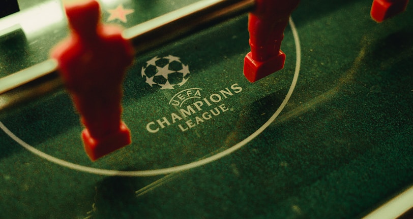A foosball table is shown with red and black player figures positioned on the rods. The surface features the UEFA Champions League logo, indicating a themed table design.