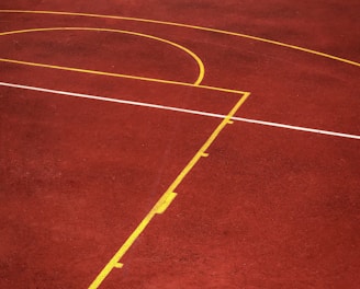 A sports court with a reddish-brown surface and bright yellow and white lines. The lines form geometric shapes, including rectangles and circles, suggesting markings for games like basketball or netball.