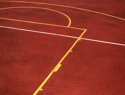 A sports court with a reddish-brown surface and bright yellow and white lines. The lines form geometric shapes, including rectangles and circles, suggesting markings for games like basketball or netball.