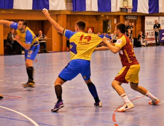 A dynamic indoor sports scene with players in motion on a court. Two athletes in blue and yellow uniforms are engaged in an intense handball game, with one player gripping the other's jersey. The background shows other players, several seated people, and advertisements on the walls.