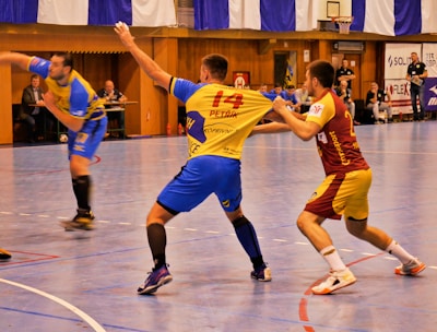 A dynamic indoor sports scene with players in motion on a court. Two athletes in blue and yellow uniforms are engaged in an intense handball game, with one player gripping the other's jersey. The background shows other players, several seated people, and advertisements on the walls.