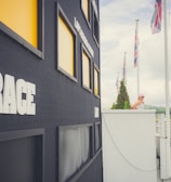 A large black scoreboard is prominently displayed with the word 'RACE' in bold white letters on its side. Surrounding the scoreboard are several yellow-highlighted rectangular slots. A woman in a white top and cap is standing on a balcony, looking towards a race track in the distance. The track is lined with green cones and several red and black flags are flying high. A vast cloudy sky and lush greenery form the backdrop.