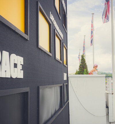 A large black scoreboard is prominently displayed with the word 'RACE' in bold white letters on its side. Surrounding the scoreboard are several yellow-highlighted rectangular slots. A woman in a white top and cap is standing on a balcony, looking towards a race track in the distance. The track is lined with green cones and several red and black flags are flying high. A vast cloudy sky and lush greenery form the backdrop.