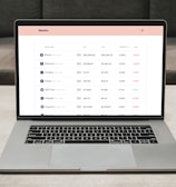 A laptop on a table displaying a cryptocurrency trading platform. The screen lists various cryptocurrencies along with their buy and sell values, spread percentages, and 24-hour change percentages. The keyboard and touchpad of the laptop are visible, set against a neutral background with a sofa in the back.