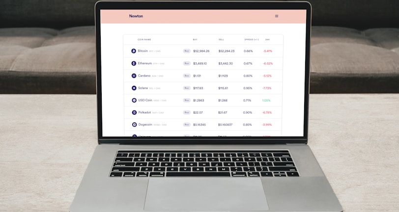 A laptop on a table displaying a cryptocurrency trading platform. The screen lists various cryptocurrencies along with their buy and sell values, spread percentages, and 24-hour change percentages. The keyboard and touchpad of the laptop are visible, set against a neutral background with a sofa in the back.