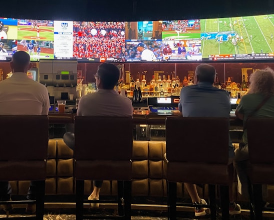A group of people sit at a bar with their backs to the camera, watching multiple sports games displayed on large screens. The bar counter is lined with various bottles of liquor, and the atmosphere suggests a casual and relaxed social setting.