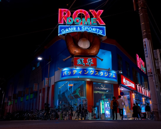 A vibrant, neon-lit facade of a gaming and sports establishment at night, featuring a large glove holding a baseball above the entrance. The building is adorned with colorful signs in both English and Japanese, with various advertisements and an illuminated sign that says 'ROX DOME'. Several people are walking towards the entrance, and bicycles are parked along the side.